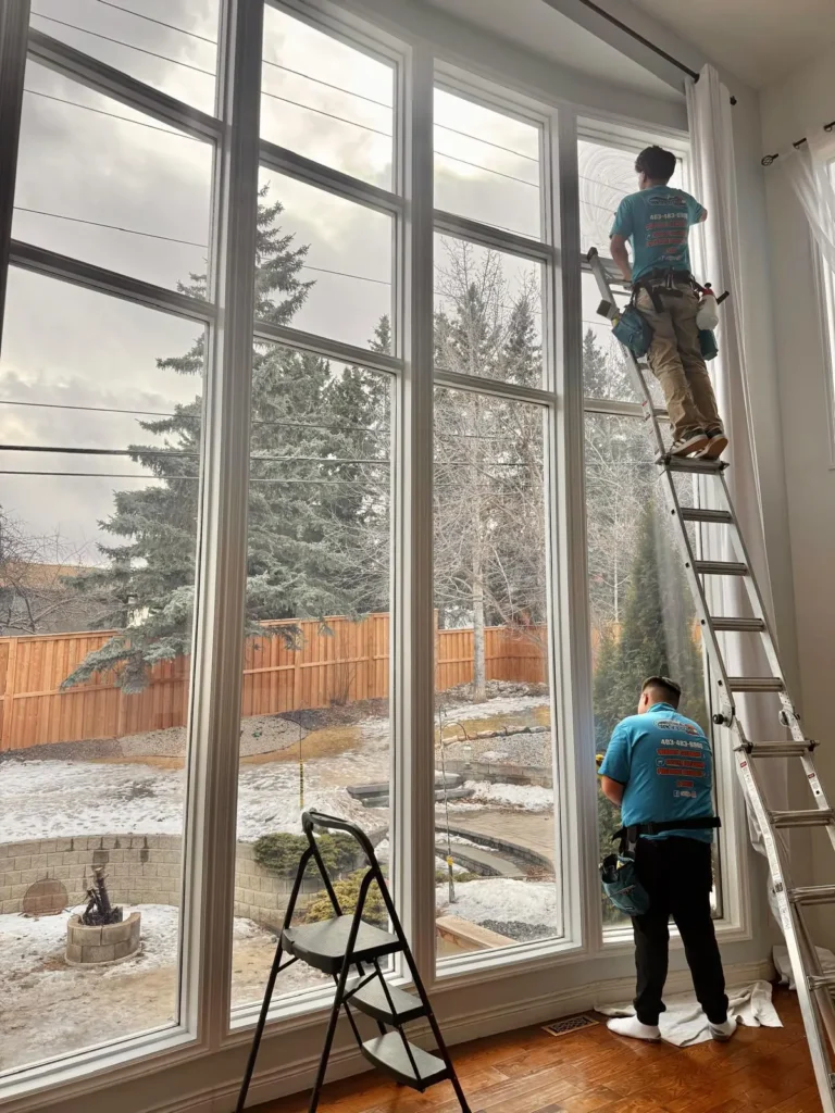 Two Pro Window Cleaning Calgary technicians cleaning tall two-storey interior windows on a ladder inside a Calgary home